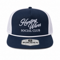 A navy blue and white trucker hat with 'Hunting Wives Social Club' embroidered on the front. The cap features a navy blue front panel and visor, white mesh back panels, and a white patch on the visor that reads 'Rhodes Headwear'.