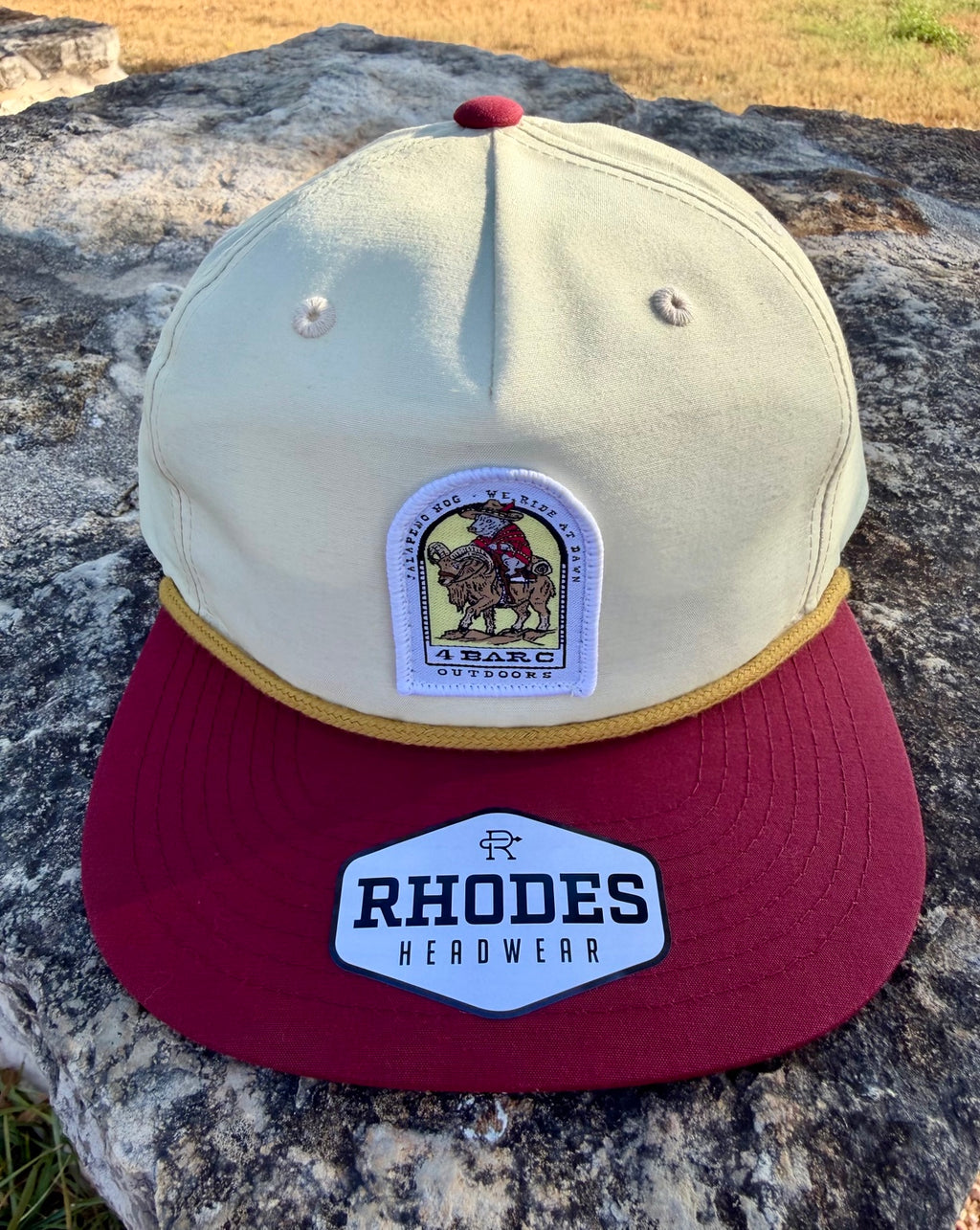 A structured baseball cap resting on a large rock outdoors. The cap has an off-white crown, a dark red flat brim, a gold piping detail around the brim base, and two embroidered patches: a central vintage-style rider patch and a white 'RHODES HEADWEAR' patch on the brim.