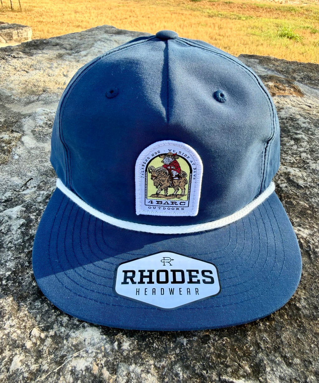 A navy blue Rhode Headwear 9FIFTY snapback cap resting on a textured stone surface outdoors. The cap features a white embroidered stripe across the front, a central arched patch with a cowboy on a horse, and a larger hexagonal patch on the brim that reads 'RHODES HEADWEAR'.